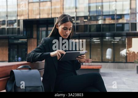 Confident businesswoman in black stylish formal attire using digital tablet. Female entrepreneur sitting outside in courtyard working remotely. Stock Photo