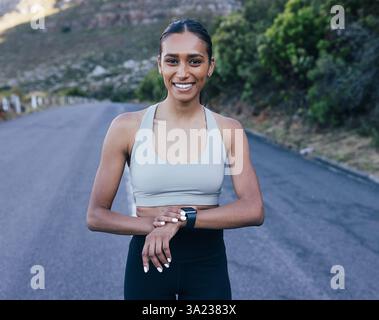 Smiling woman in fitness attire looking at camera and using smartwatches while standing on abandoned road Stock Photo