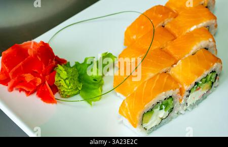 Fresh sushi rolls are arranged neatly on a white plate. The dish features vibrant salmon, crunchy vegetables, and is accompanied by pickled ginger and Stock Photo