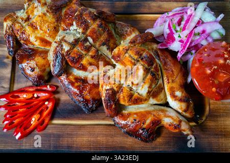 Three pieces of grilled chicken with a crispy skin are arranged on a wooden platter. Accompanied by fresh sliced onions, tomatoes, and red chilies for Stock Photo