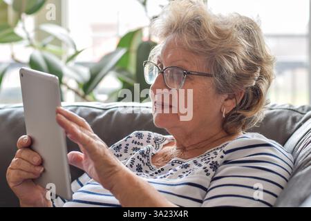 Senior woman sitting on couch at home using wireless tablet Stock Photo