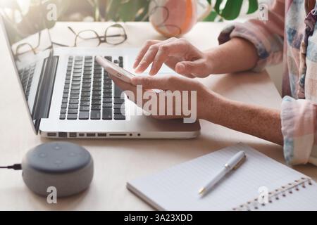 Hands of woman sitting at desk in front of laptop computer using mobile phone, side view Stock Photo