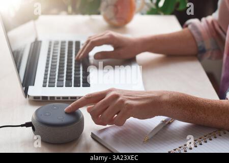 Woman sitting at desk typing on laptop computer and pressing button on virtual assistant smart speaker Stock Photo