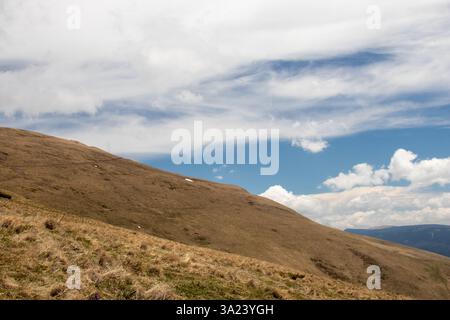 A vast hillside features dry grass and patches of snow under a bright blue sky. Fluffy white clouds float above, creating a serene atmosphere perfect Stock Photo