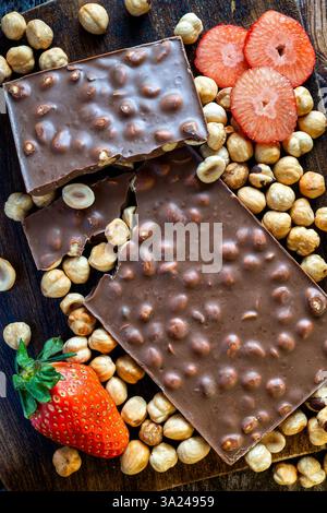 Pieces of chocolate bar with hazelnuts on a dark wooden board. At the bottom, a whole red strawberry and above, three pieces of fresh strawberries, pr Stock Photo