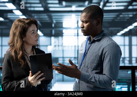 Computer scientists in data center using tablet, analyzing metrics, optimizing storage volumes. Server farm coworkers using device, doing brainstorming, ensuring seamless operations Stock Photo