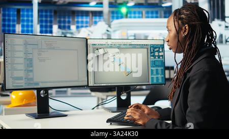 Programmer in photovoltaics factory using PC to update software of robot arms placing solar panels on assembly lines. Woman coding on computer, upgrading solar plant machinery, camera A Stock Photo