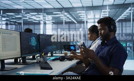 Admin in server room using mobile phone, relaxing after ensuring minimal downtime. Data center IT expert texting on smartphone, taking break from monitoring performance, camera A Stock Photo