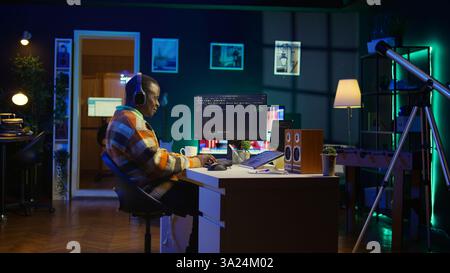 Black programmer remotely working in home office writing code on computer at his desk. Programming engineer IT expert in apartment typing lines of code at workstation, teleworking. Camera B. Stock Photo