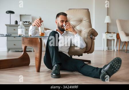 Sitting on the floor, sad. Upset young man drinker alcoholic is indoors. Stock Photo
