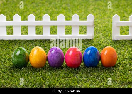 Multicolored Easter eggs stand in a row on a green lawn against a white wooden fence Stock Photo