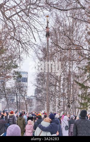 Yekaterinburg, Russia - February 26, 2025: Young man climbs tall, smooth wooden pole to reach prize during traditional Maslenitsa celebration in city Stock Photo