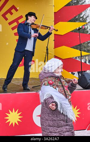 Yekaterinburg, Russia - February 26, 2025: Elderly woman in traditional shawl dances at Maslenitsa festival. Violinist in elegant suit plays on stage Stock Photo