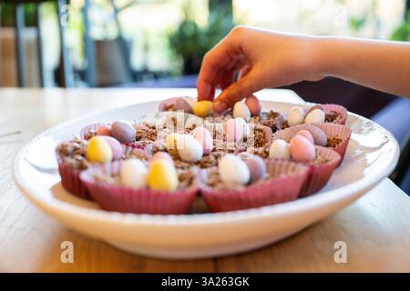 Hand putting decorations on to Easter cup cakes in dish on kitchen table. Stock Photo