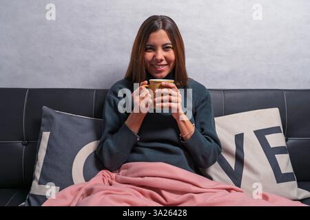 Woman enjoying a cozy moment on the sofa, wrapped in a blanket and holding a warm beverage Stock Photo