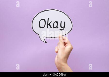 Woman holding paper speech bubble with word Okay on lilac background, closeup Stock Photo