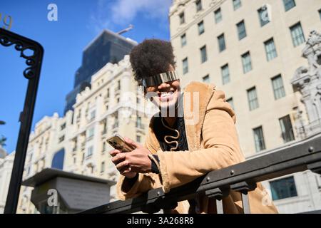 Smiling androgynous person with futuristic sunglasses using phone and leaning on a railing in a city Stock Photo