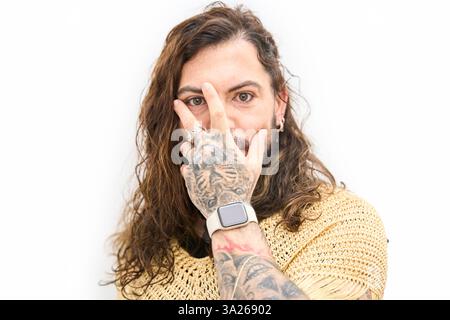 Portrait of a young non binary latin influencer with long hair, beard and tattoos covering face with hand on white background Stock Photo