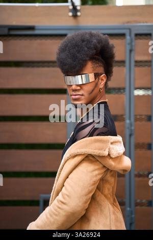 Androgynous person with afro hair wearing futuristic glasses and a brown coat is posing in front of a wooden gate Stock Photo