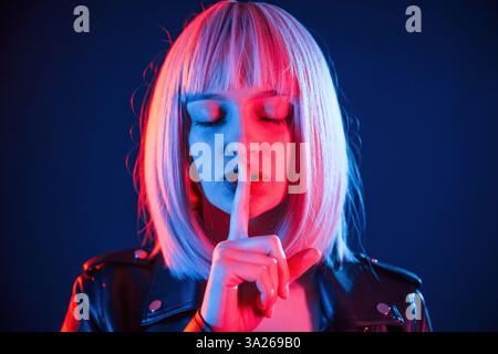 Finger by the lips, silence. Woman with white hair is in studio with neon colors. Stock Photo