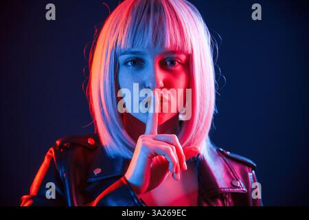 Finger by the lips, silence. Woman with white hair is in studio with neon colors. Stock Photo