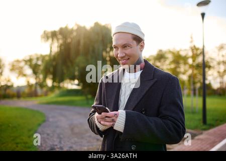 Handsome young man with tattoos smiles while texting, dressed in modern, chic attire during sunset. Stock Photo