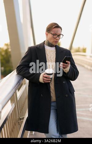 Stylish man with tattoos sips coffee while intently checking his phone. Stock Photo