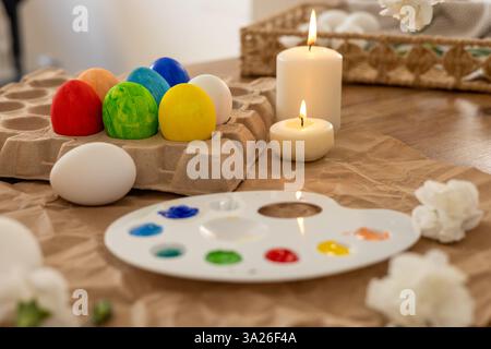 Brightly colored Easter eggs are arranged on a table, accompanied by a palette of paints and candles. White flowers and a spring atmosphere enhance th Stock Photo