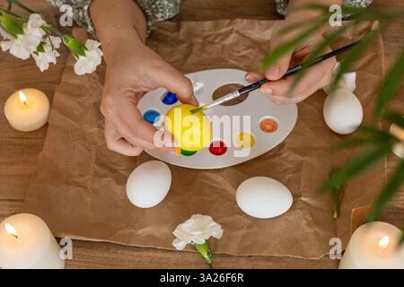 Hands carefully paint white eggs using a palette of vibrant colors surrounded by candles and flowers. This activity captures the festive spirit of Eas Stock Photo
