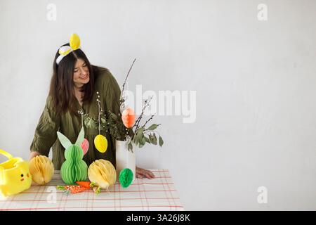 Happy Easter! Stylish woman in bunny ears decorating with colorful easter paper eggs, bunny and carrots on table. Easter table setting and Easter hunt Stock Photo