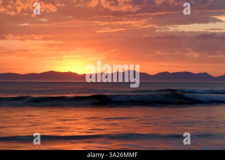 Wilson's Promontory seen from across the sea with purple and orange hues of the rising sun reflect in the glassy waves breaking on the shore. Stock Photo