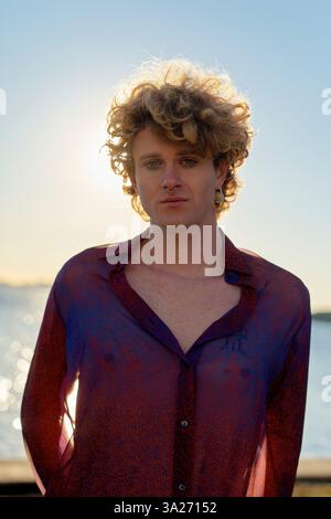 Person with curly hair wearing a sheer shirt stands by water with the sun setting behind them. Miami, FL, USA Stock Photo