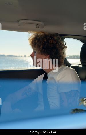 Young person with curly hair and tattoos looking out a car window at a scenic ocean view. Miami, FL, USA Stock Photo
