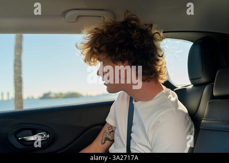 Young man with curly hair looks out car window toward the ocean on a sunny day. Miami, FL, USA Stock Photo