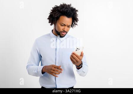 Disappointed African-American man watching on the smartphone standing isolated on white background, black guy receved bad news, surprised by what he sees on phone screen, negative emotions Stock Photo