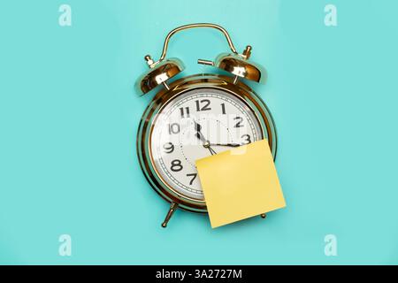 A blank yellow sticky note glued on the face of a bell alarm clock on a light blue background with copy space Stock Photo