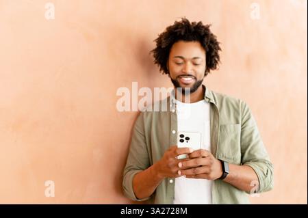 Studio shot of young African American man standing isolated on wall holding mobile phone and typing message, chatting with friend on social media online, using dating app, enjoying new application Stock Photo