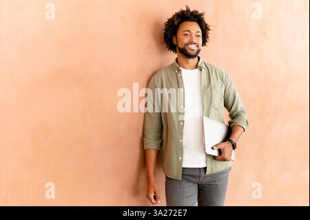 Portrait of young african american guy standing isolated on wall holding laptop, looking distance, going on work, showing positive emotions. Technology and people concept Stock Photo