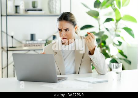 Irritated caucasian businesswoman looking focused and determined while working on her laptop in the office, showing a sense of frustration and determination to resolve an issue, looking dessatisfied Stock Photo