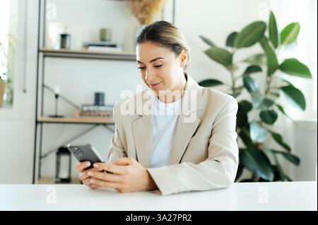 Positive focused caucasian woman in a beige blazer uses her smartphone while working, checking messages, responding to emails, or managing business tasks with efficiency sitting at a desk in an office Stock Photo