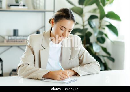 Focused caucasian businesswoman in a beige blazer writes notes in a notebook while working on important tasks, planning projects, or organizing ideas with dedication and concentration Stock Photo