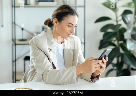Focused busy caucasian businesswoman in a beige blazer sits at a modern office desk using her smartphone, checking important information or responding to a message with a concentrated expression Stock Photo