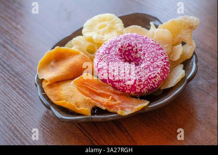 A delightful arrangement of a pink frosted donut, dried mango slices, pineapple rings, and crispy snacks sits on a wooden table, inviting indulgence a Stock Photo