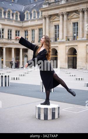 A fashionable woman in a black outfit gracefully balances on a striped column at Palais-Royal in Paris, striking a poised ballet-inspired pose amid classic architecture.  Stock Photo
