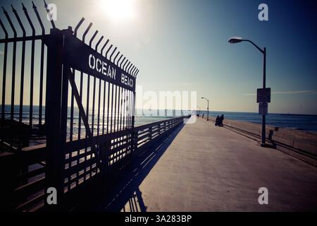 A scenic view of a pier leading to the ocean, with a gate labeled 'OCEAN BEACH'. The sun shines brightly in the sky, casting reflections on the water. Stock Photo
