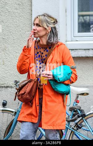 Young woman with long blonde hair wearing a bright orange jacket standing in street and smoking - Tours, Indre-et-Loire (37), France. Stock Photo