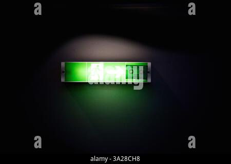 A glowing green exit sign with an arrow pointing to the right, set against a dark background. The sign features a figure walking and is illuminated fo Stock Photo