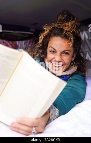 smiling and curly-haired woman looking to camera showing a book in her camper on sant jordi Stock Photo
