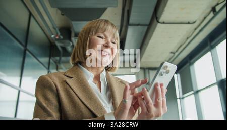 A mature businesswoman using a smartphone in a modern office environment, focusing on professional tasks or communication, highlighting confidence and Stock Photo