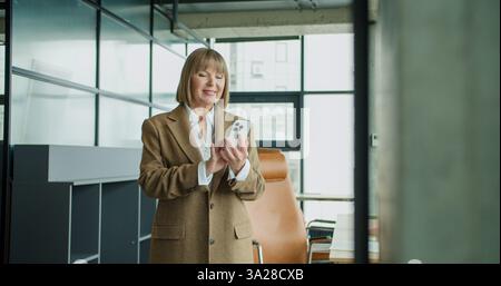 A mature businesswoman using a smartphone in a modern office setting, checking messages or emails while standing confidently, showcasing Stock Photo